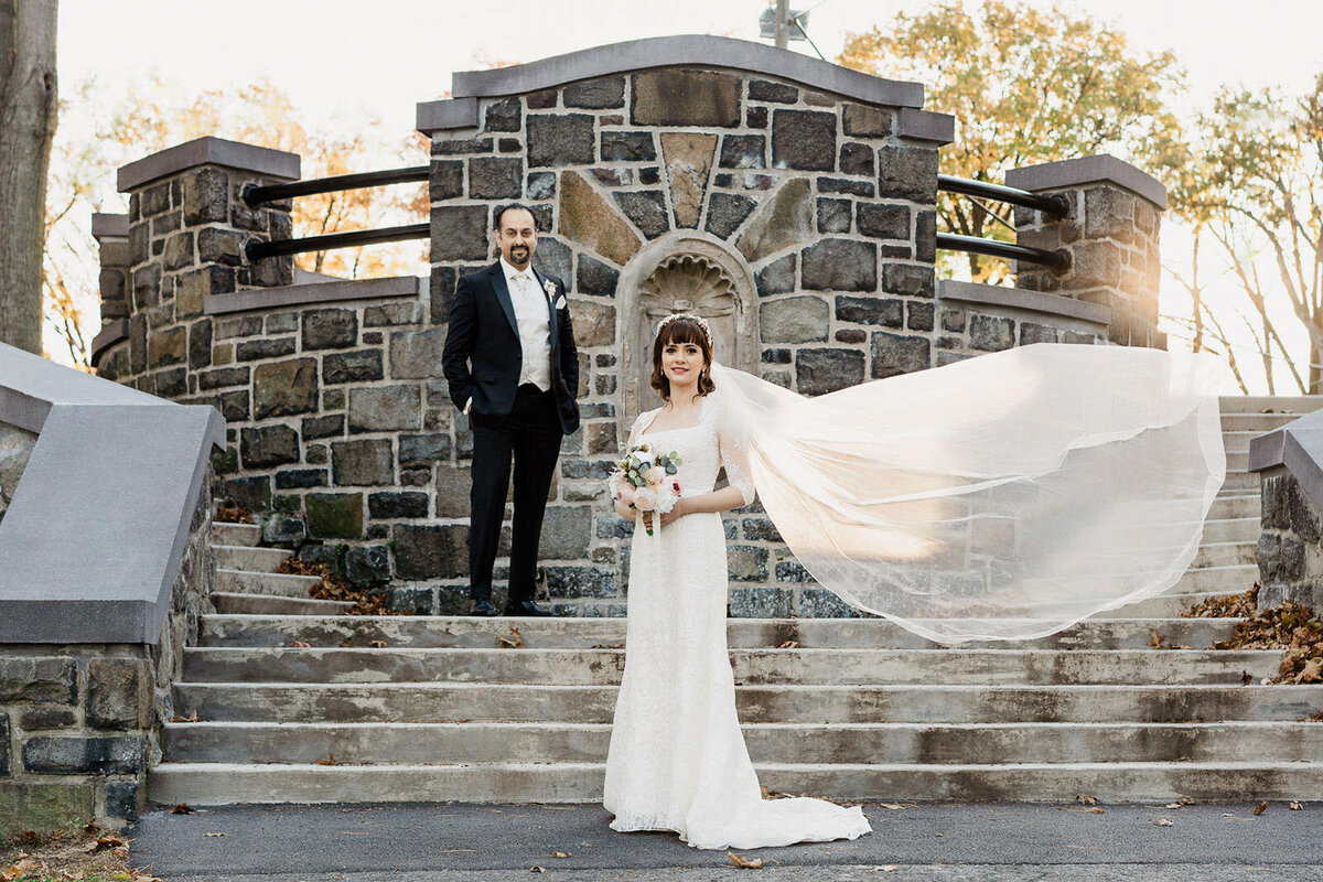 Bride and groom standing on a stone staircase with the bride’s veil blowing in the wind during their New Jersey wedding portrait.
