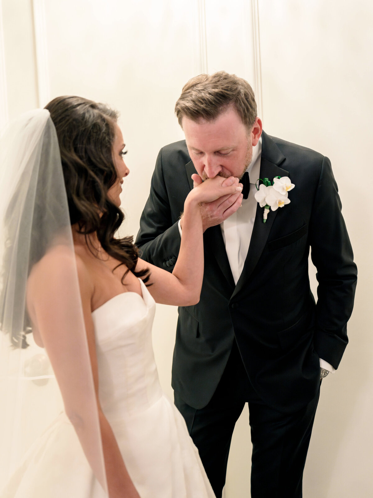 A bride in a white strapless gown and veil stands as a groom in a black suit and bow tie gently kisses her hand. Both are in an elegant setting, sharing an intimate moment.
