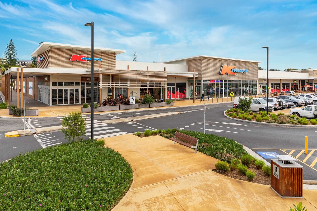 A wide-angle view of the Marina Shopping Centre in Port Macquarie, showing the clean modern façade of the building with landscaped gardens and footpath leading to the entrance.