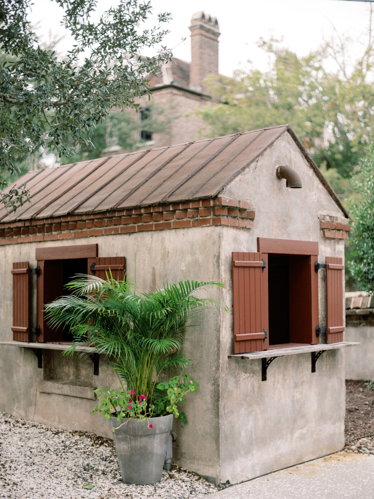 This quaint rustic building at Twenty-One Magazine features a stone facade with decorative brick and wooden shutters, surrounded by vibrant greenery, creating a peaceful outdoor atmosphere.
