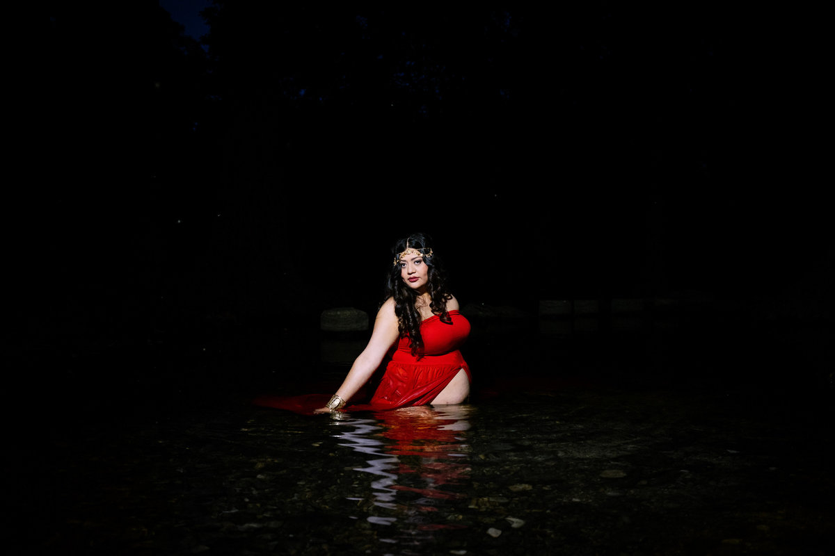 Pregnant woman in red maternity dress sitting in river water in Cibolo Nature Center.