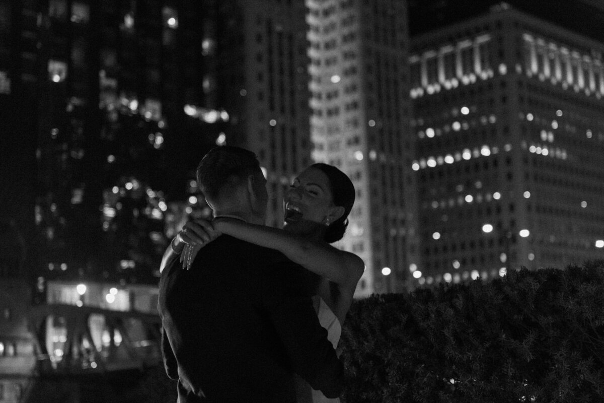 A moody and cinematic black and white wedding photo of a bride and groom embracing outside at night along the Chicago Riverwalk.