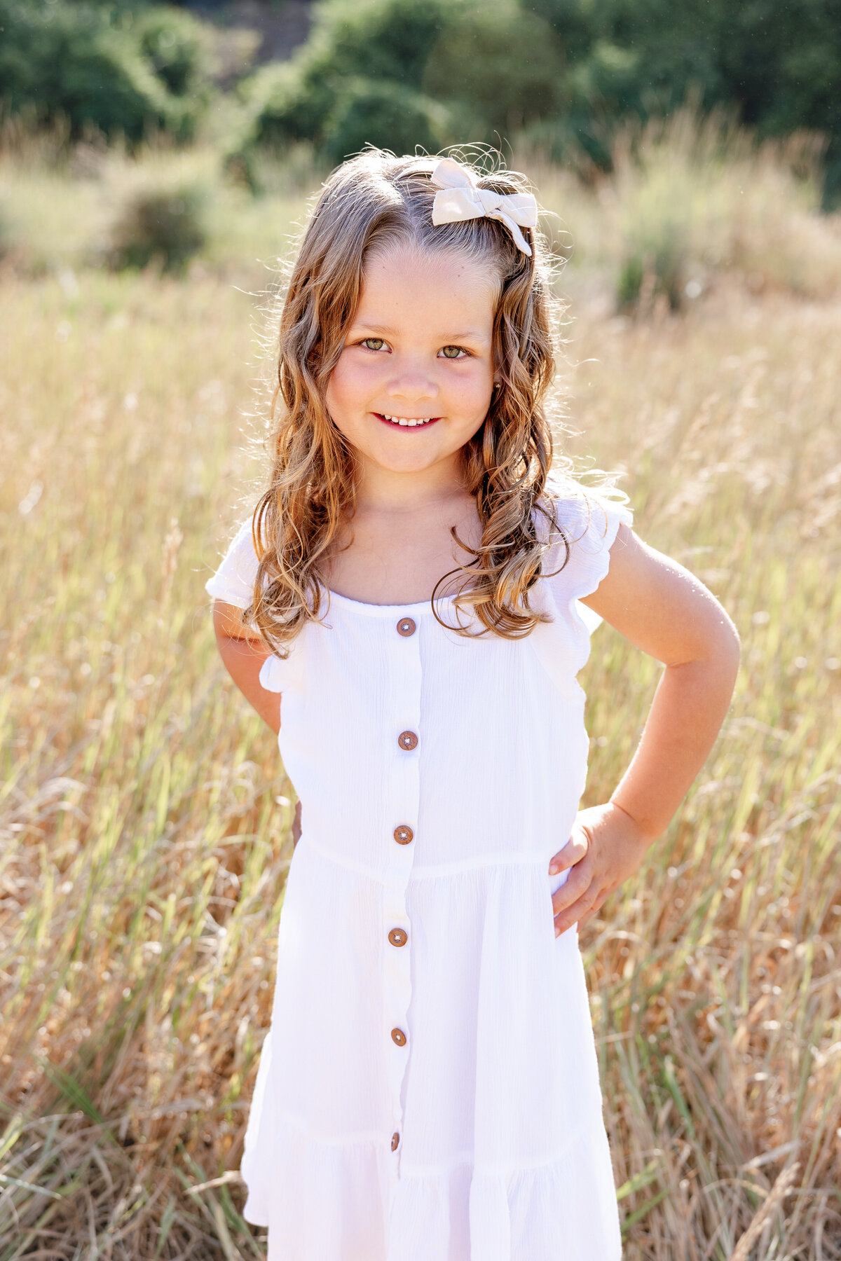 A young girl in a white dress stands in a golden field, smiling brightly. Her curly hair, adorned with a bow, catches the sunlight as she radiates happiness in this natural setting