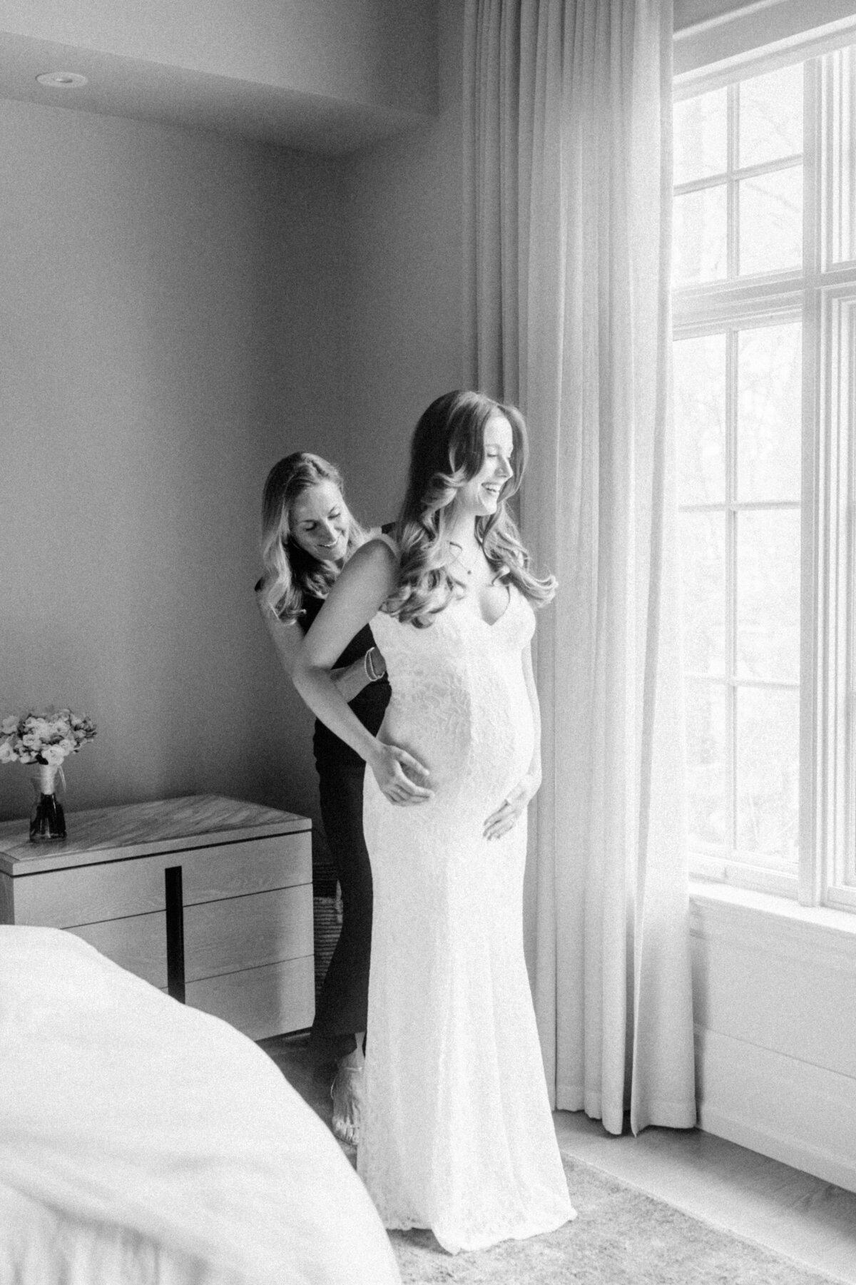 A serene, black-and-white image of a mother assisting her pregnant daughter in adjusting her lace bridal gown, capturing a precious moment of love and support