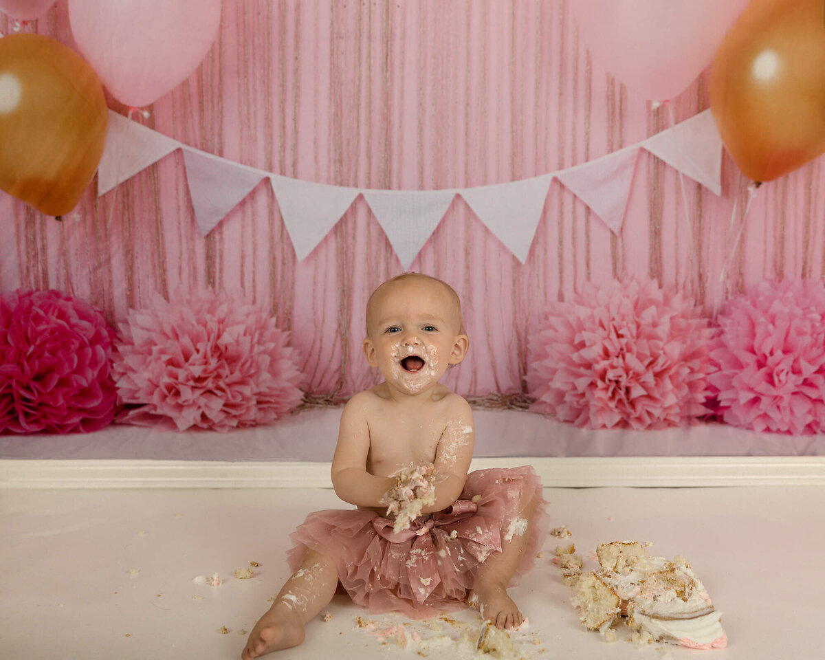 One year old girl making a mess during her cake smash. Backgound is pink design with balloons.