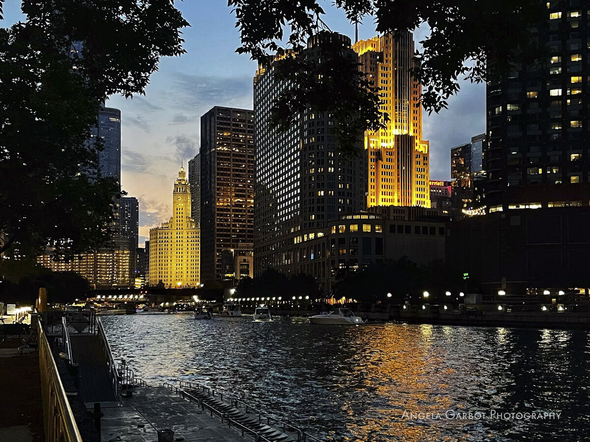 Chicago Riverwalk at sunset on the bank of the Chicago River looking west with trees, boats and the Wrigley Building in full view.