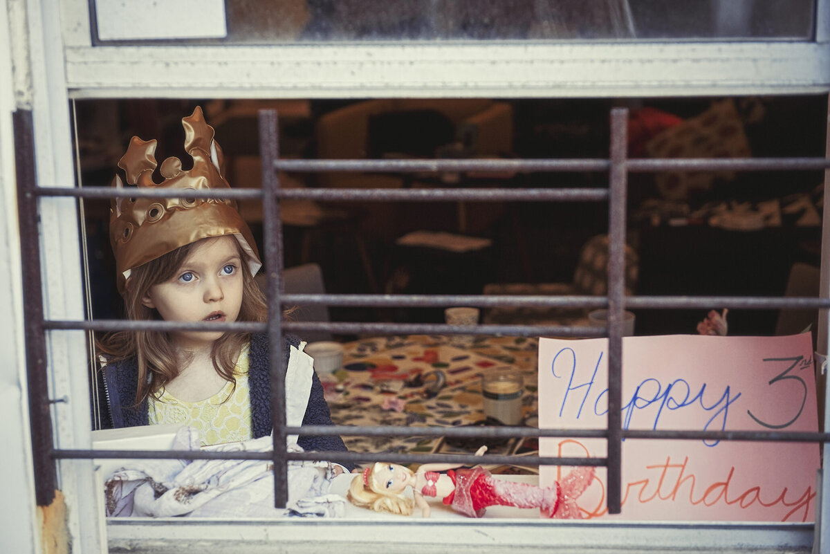 A child looking out a window with  paper next to her in the window that says happy birthday