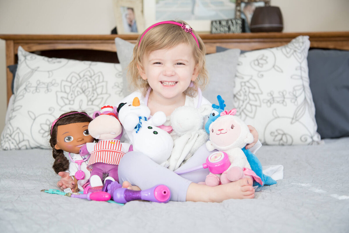 blonde toddler girl smiling with her favorite stuffies on a bed to celebrate her 3  year milestone