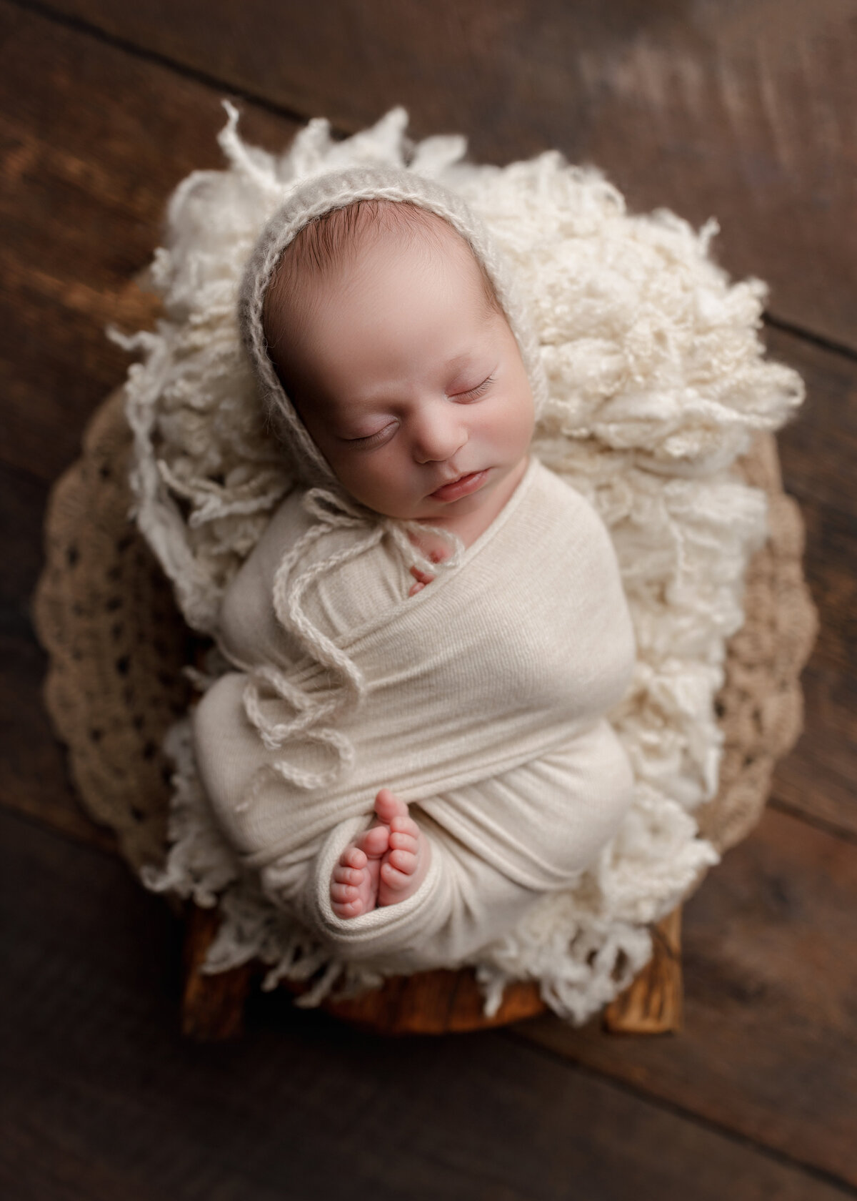 Baby girl newborn photos from West Palm Beach, FL photographer. Baby girl is swaddled in a cream knit wrap with her toes peeking out of the bottom. Baby is sleeping in a basket covered with cream knit. Baby is wearing a knit bonnet. Aerial image.