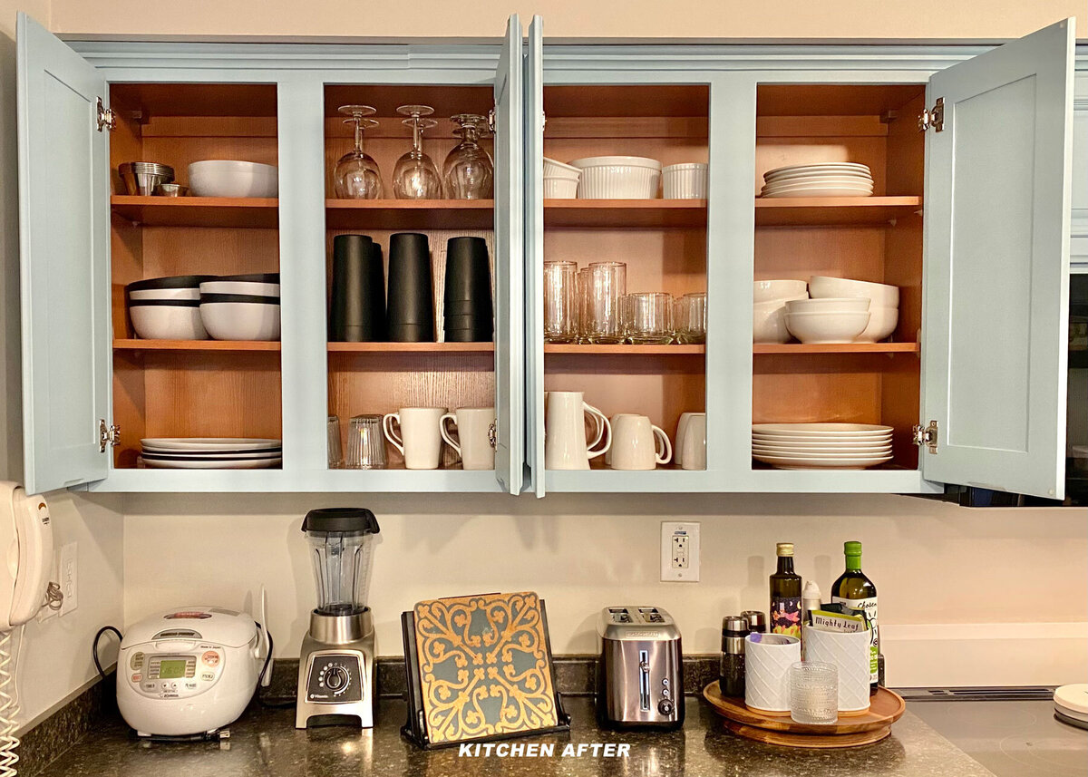 Open kitchen cabinets with neatly arranged dishes, glasses, and mugs. Below, countertop appliances include a rice cooker and blender. A tray holds olive oil and other items. Cabinets are light blue, and text reads "KITCHEN AFTER.