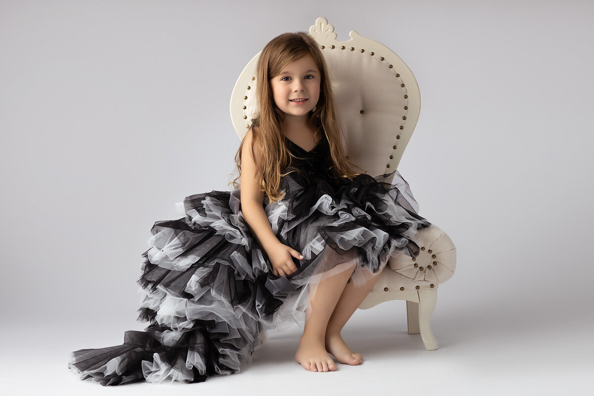 Dallas family photographer captures a little girl in a black and white ruffled gown, sitting on a vintage chair, barefoot.