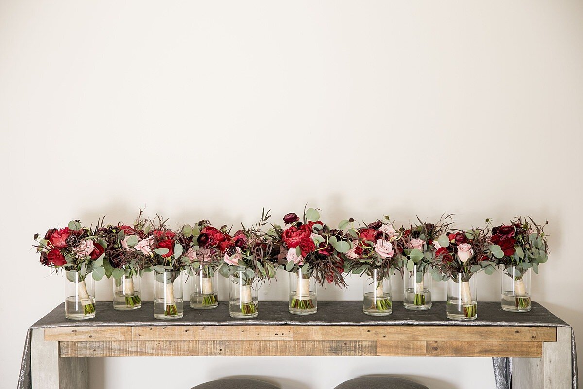 Twelve bridesmaid bouquets in glass vases sitting on long wooden table against a white wall. The bouquets are red, blush and burgundy with hints of purple accente with greenery and tied with ivory satin ribbon.