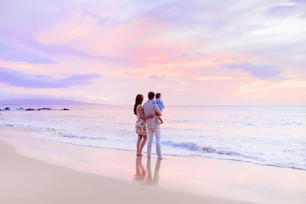 Mom in cute floral dress holds her husband just after sunrise as their family portraits are captured on Maui by Love + Water Photography