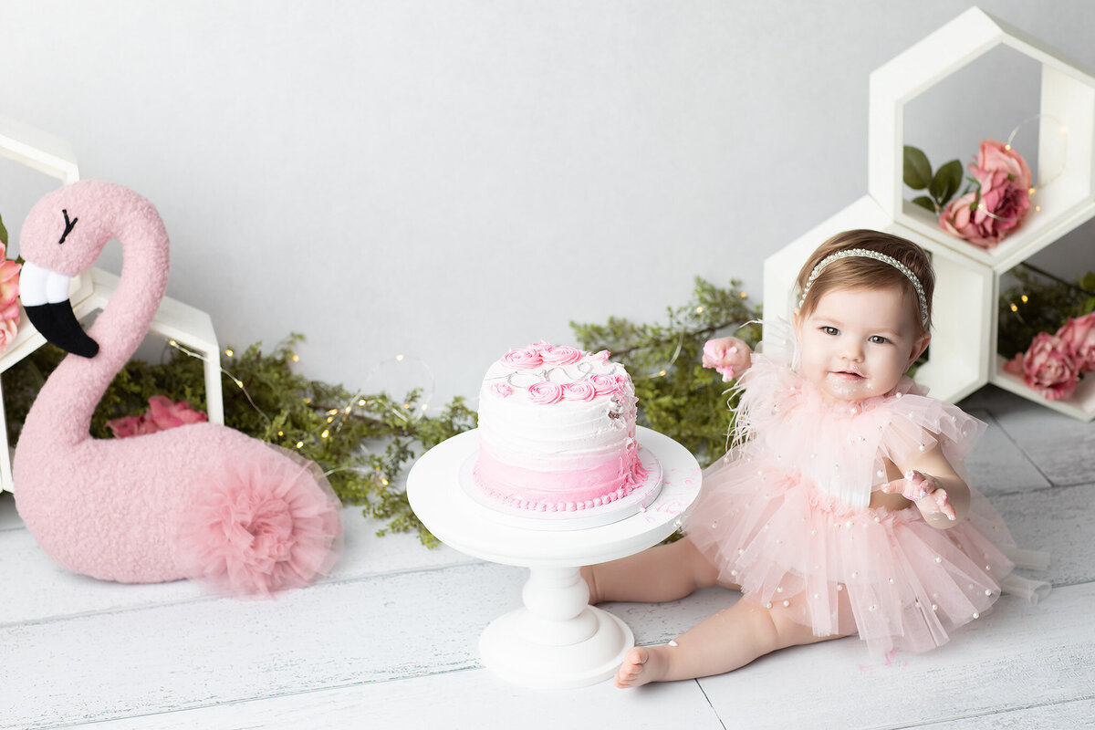 Dallas baby photographer captures a sweet moment of a baby girl in a pearl-accented pink dress enjoying her pink ombré cake during a first birthday milestone session with soft florals and greenery in the background.