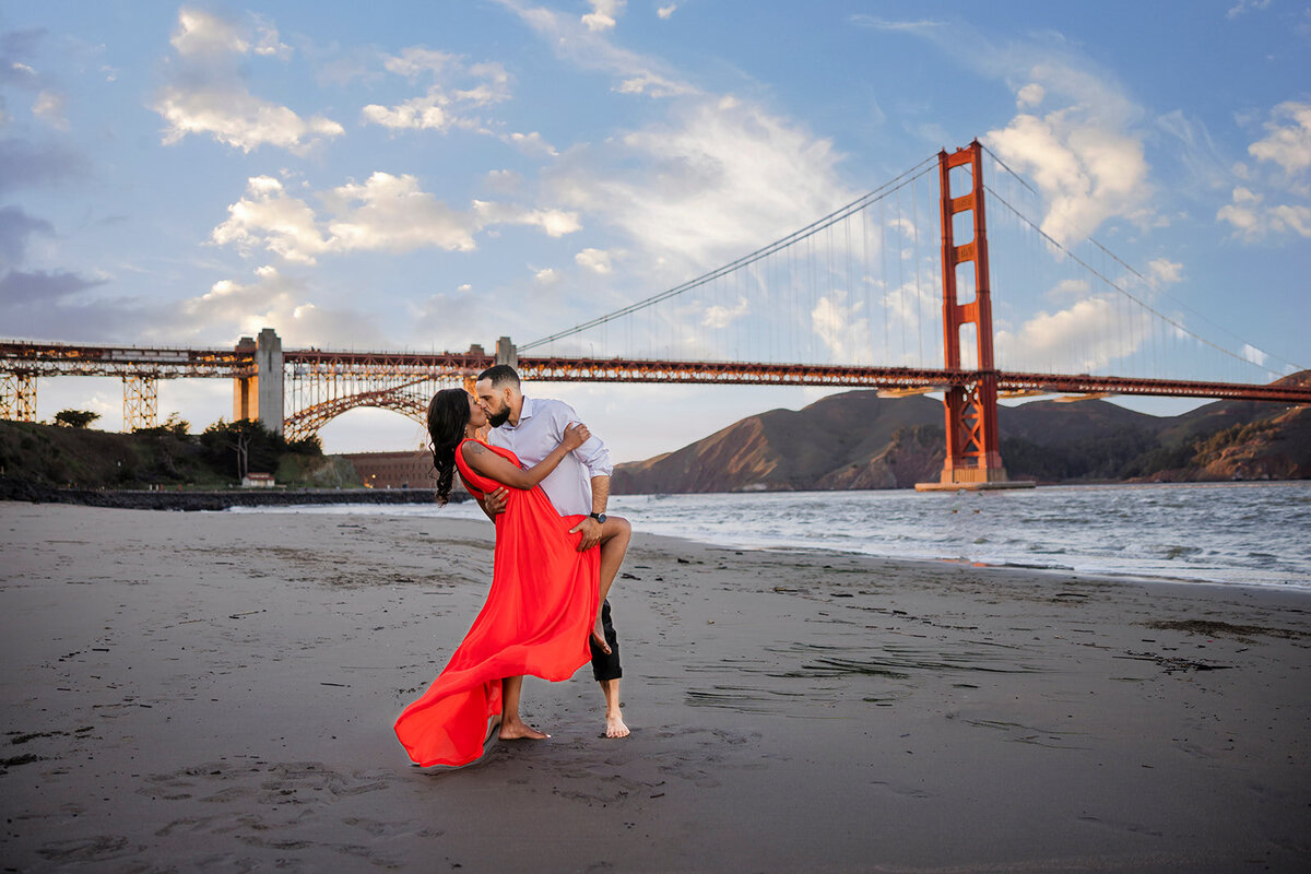 During an engagement session on the beach at the bottom of the golden gate bridge a man dips his fiance who is wearing a red dress and goes in for a kiss.  Photo creatively captured and edited by philippe studio pro, a wedding photographer studio in Sacramento, CA.