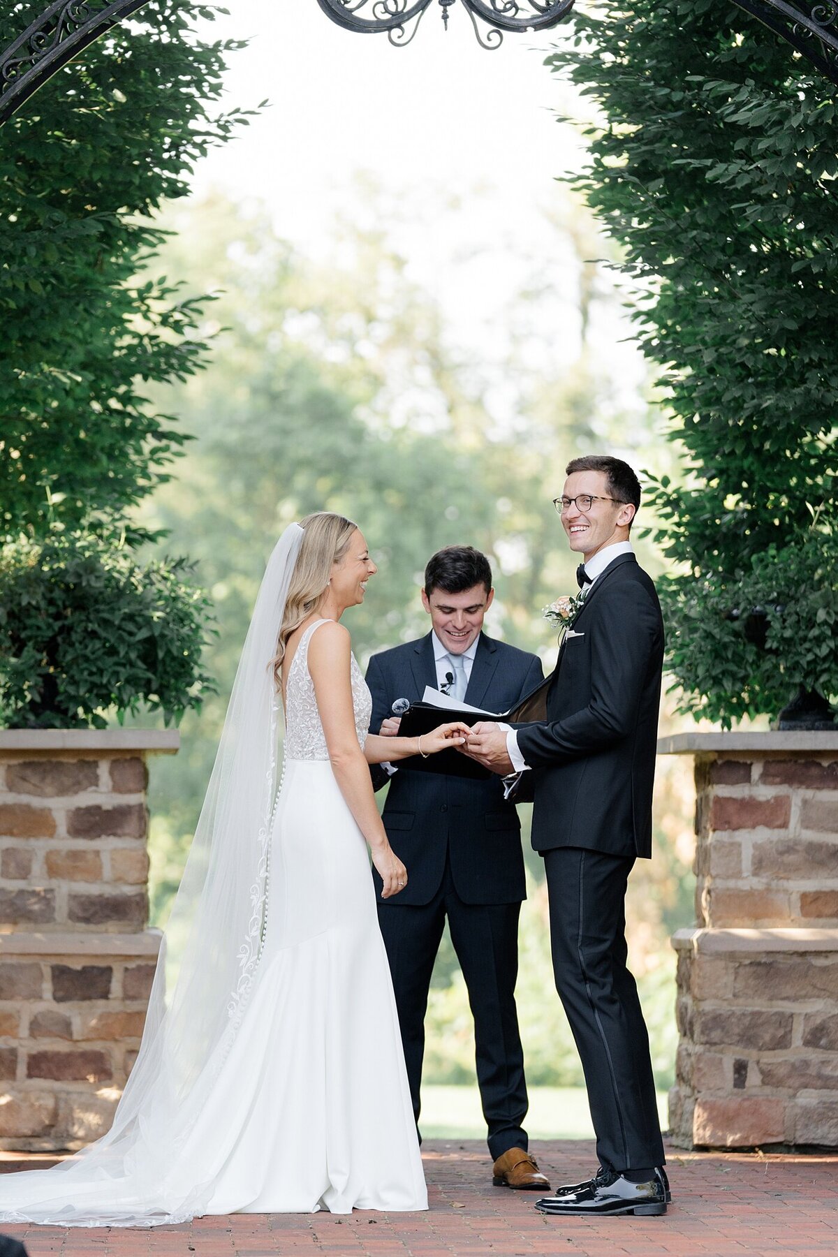 Columbus wedding  photographer captures outdoor wedding ceremony at the Pinnacle with groom putting on a ring for the bride with shrubs and brick pillar behind them