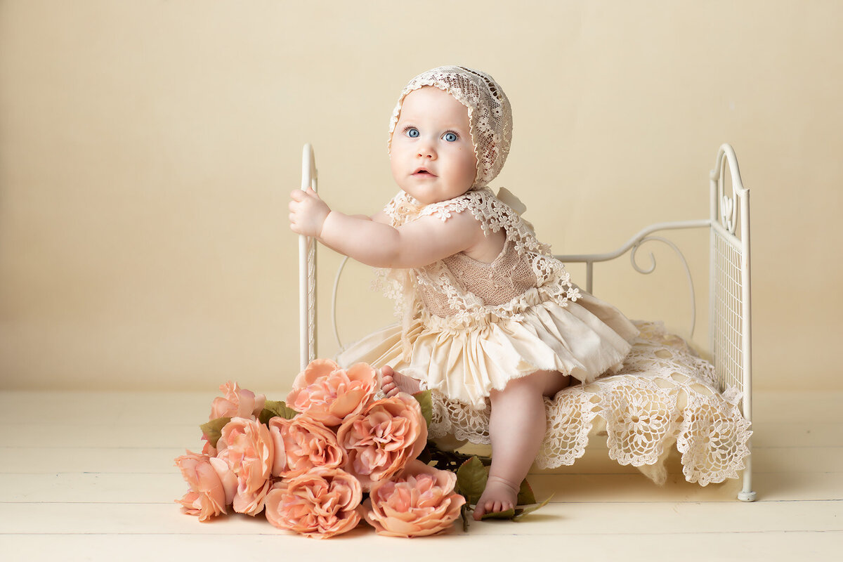A baby girl dressed in delicate lace attire and bonnet sits on an ornate vintage-style metal bed surrounded by blush peach roses. The soft, cream-toned background enhances the elegant scene, captured by a Dallas baby photographer specializing in milestone photography.