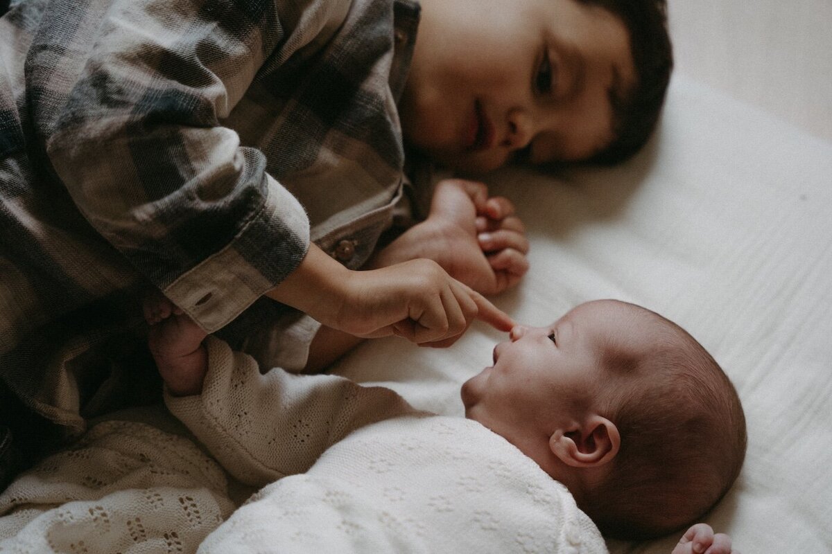 Le grand frère touche du bout du doigt le nez du bébé allongé à côté de lui. Par Kamescop, photographe famille à Nantes.