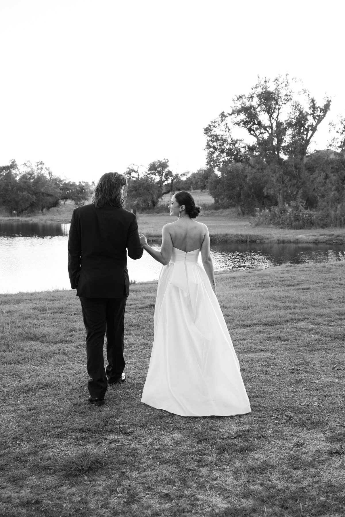 An Austin wedding photographer captures the serene moment of a bride and groom walking in front of a picturesque pond.