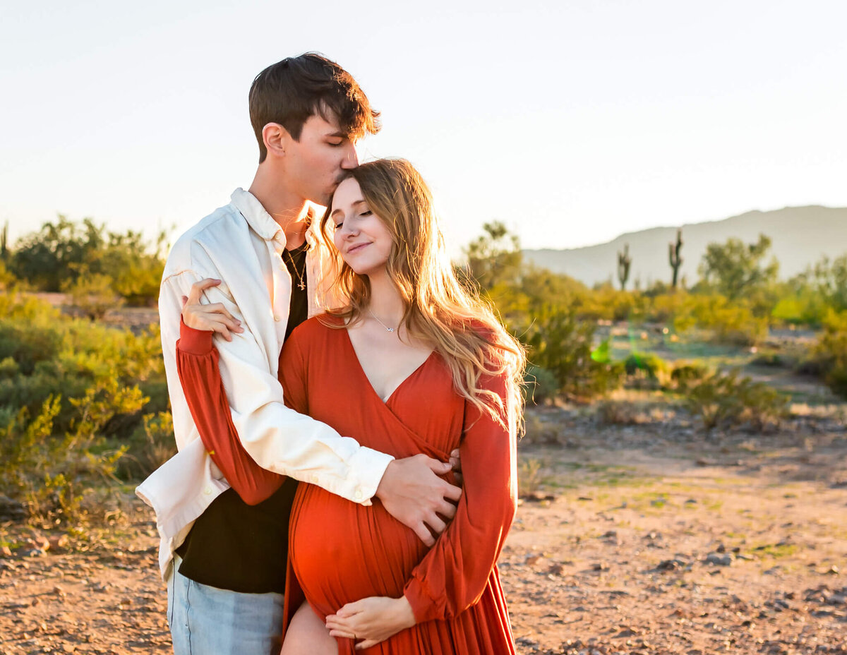 young father kissing expecting wife on top of her head while she holds her baby bump