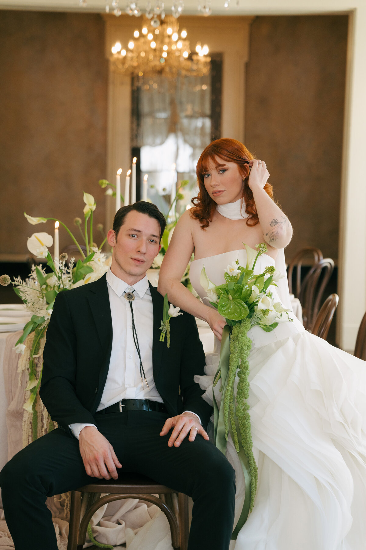 A red-headed bride holding a green floral bouquet leaned up against a groom sitting in a chair looking at the camera at Riverwood Mansion in Tennessee. 