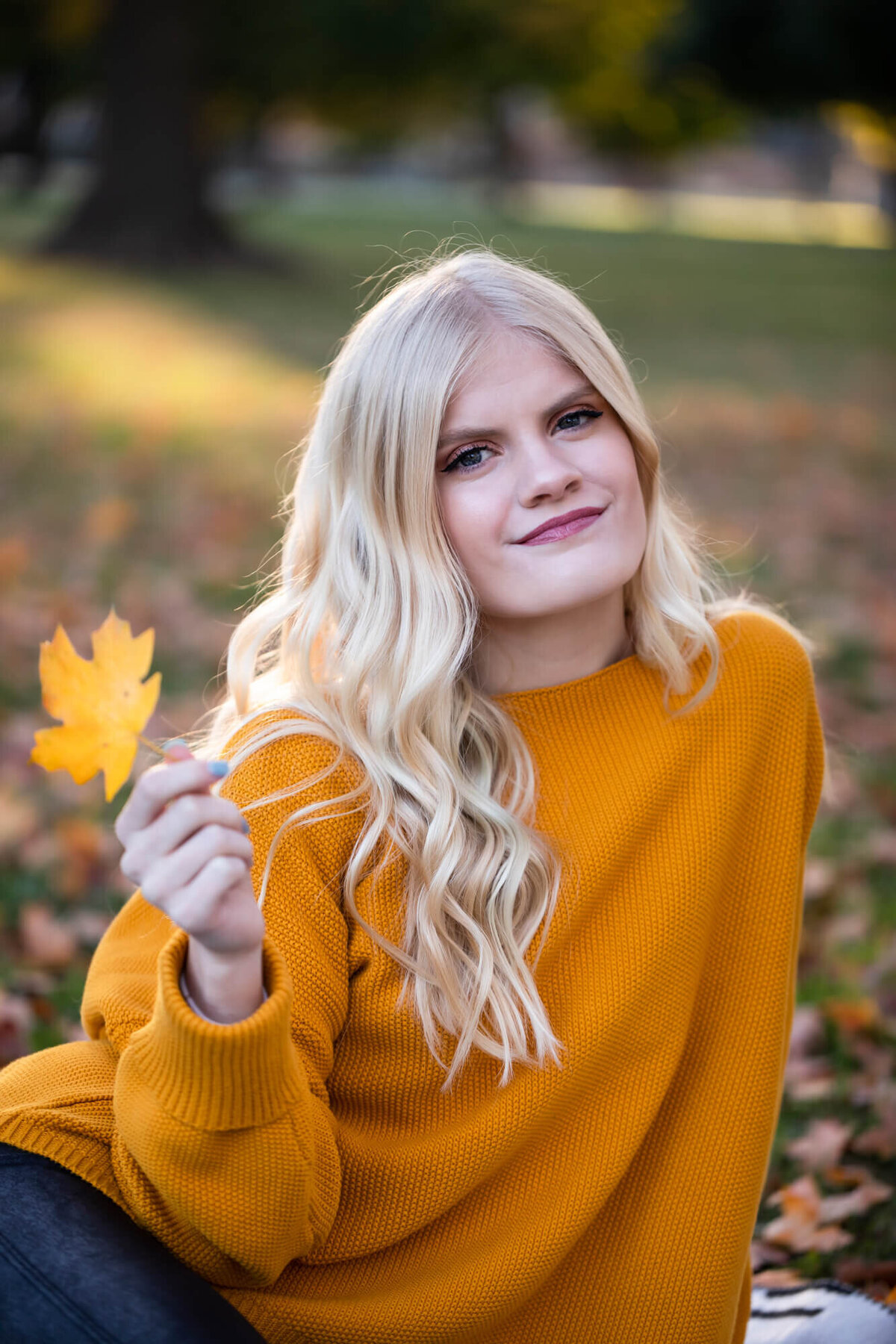 A lovely blonde girl poses for a Fall senior portrait wearing a dark yellow sweater and twirling a golden maple leaf. Captured by Springfield, MO senior photographer Dynae Levingston.