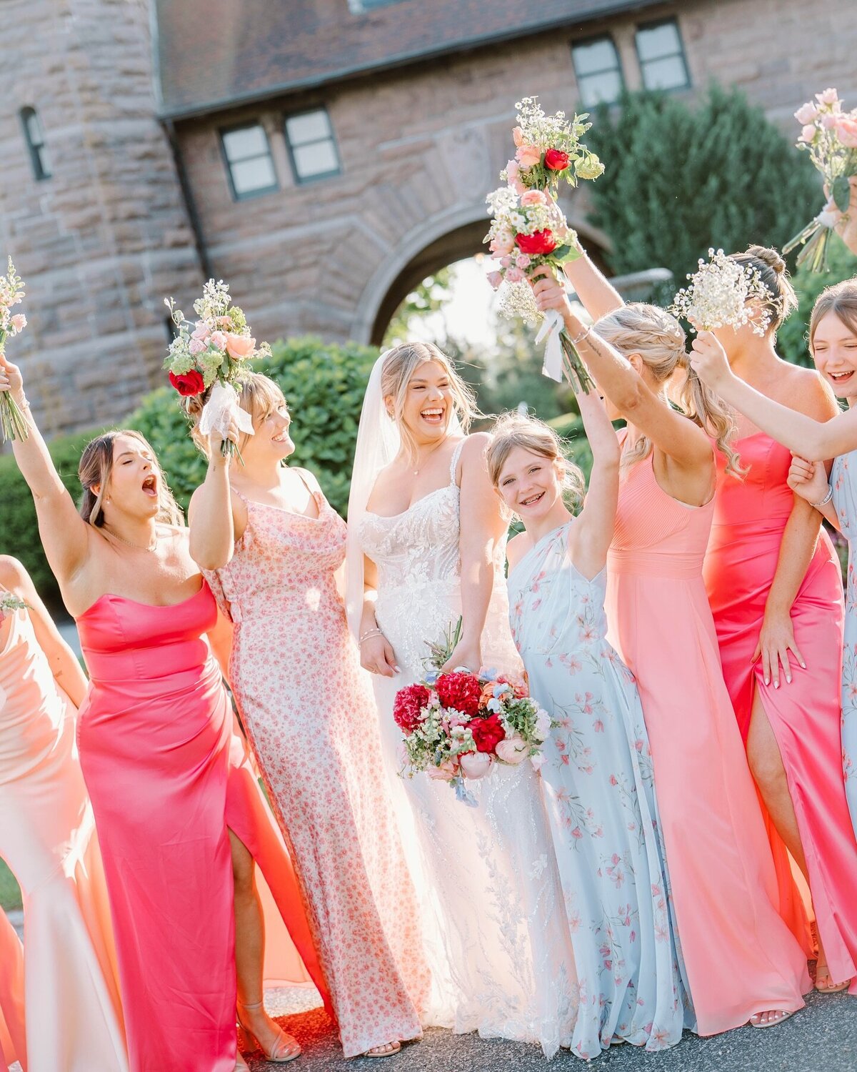 Wedding Photographer RI | A joyful bride in a white gown and veil stands with bridesmaids in pink and floral dresses, holding bouquets aloft in celebration, against a stone archway backdrop.