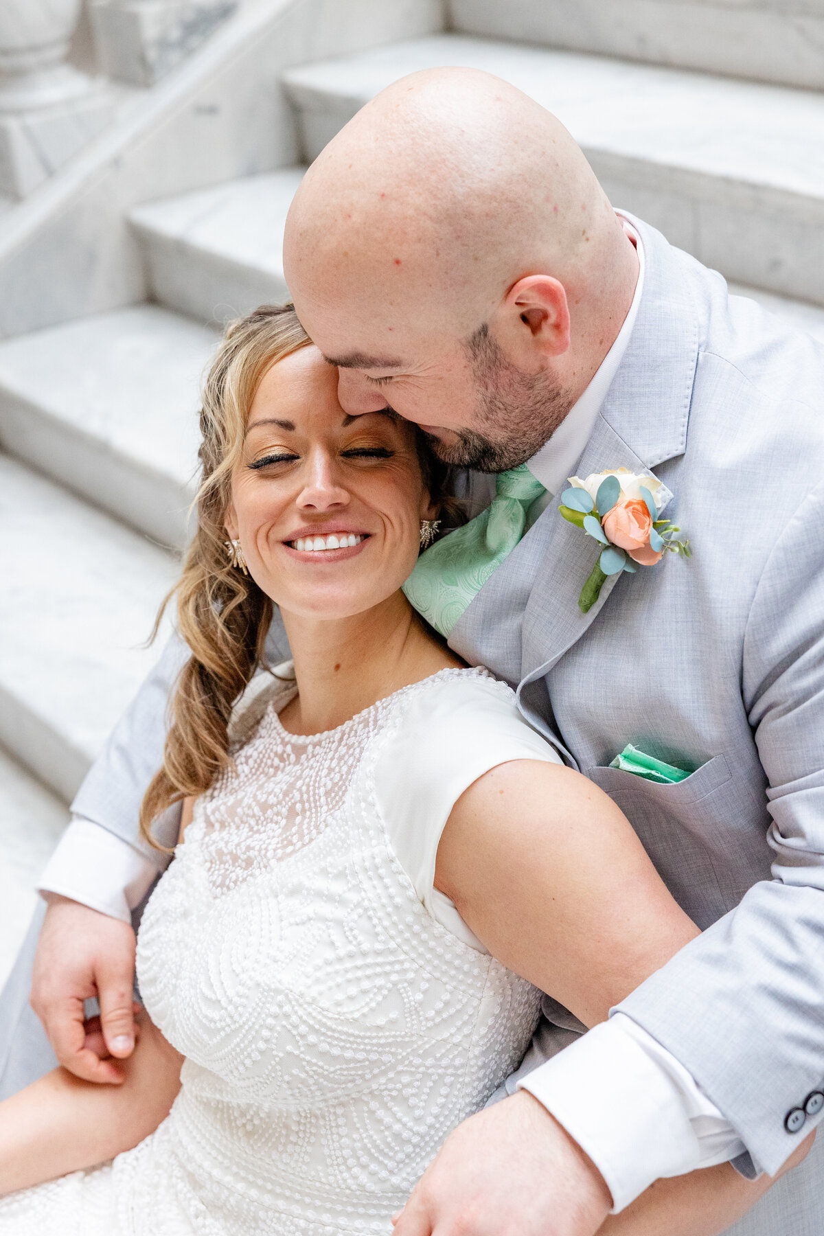 A groom kisses his bride’s forehead as she smiles softly, leaning into him on the marble steps of the Utah State Capitol.