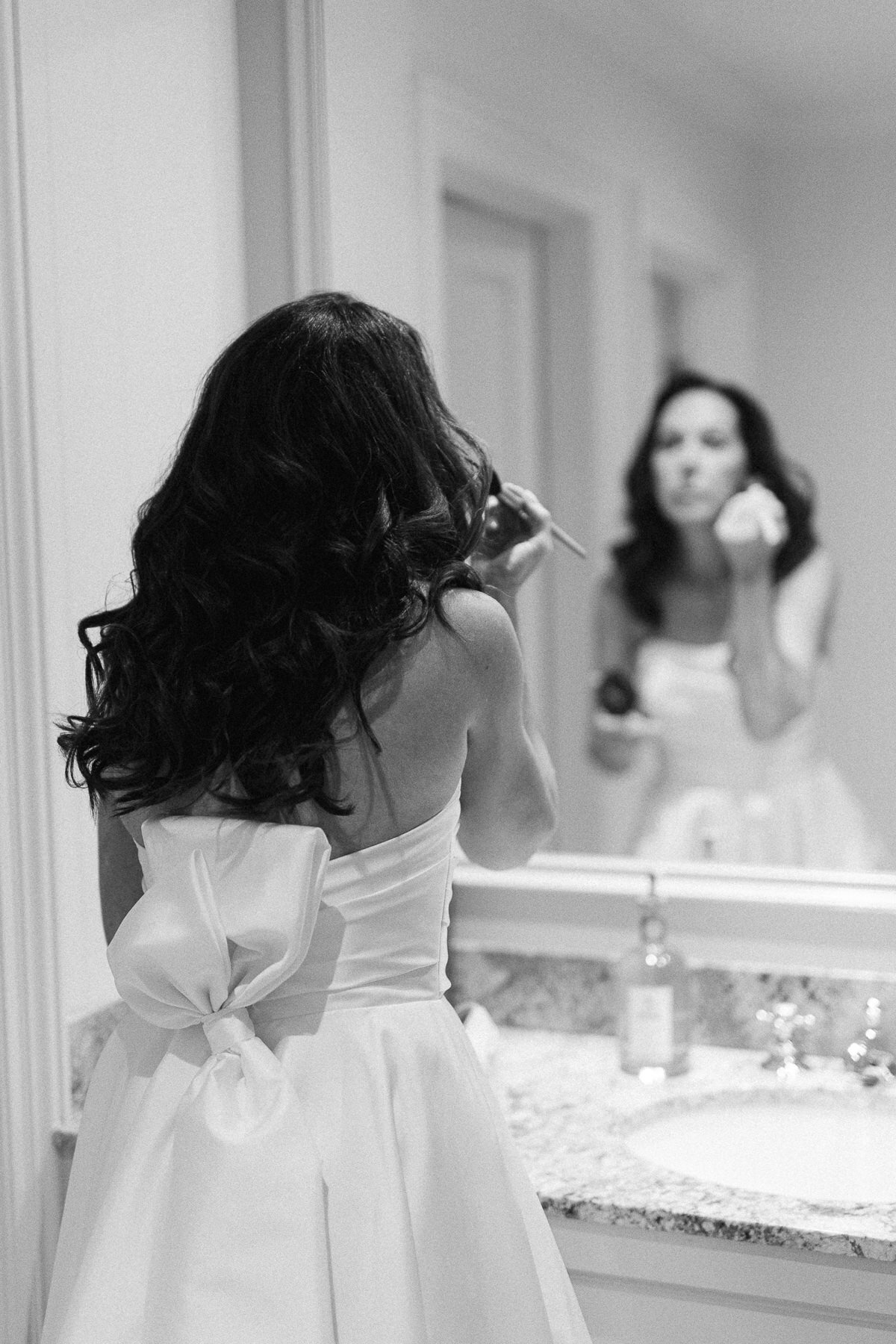 Une mariée de maquille et se regarde devant le miroir avec sa robe de mariage qui a une grande boucle dans le dos.