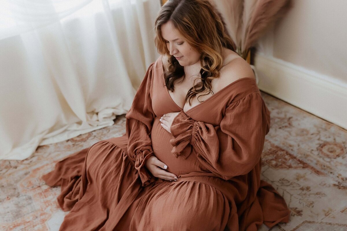 Pregnant woman in a brown dress sits on a patterned rug, gently holding her belly, with light streaming through a window draped in white curtains.
