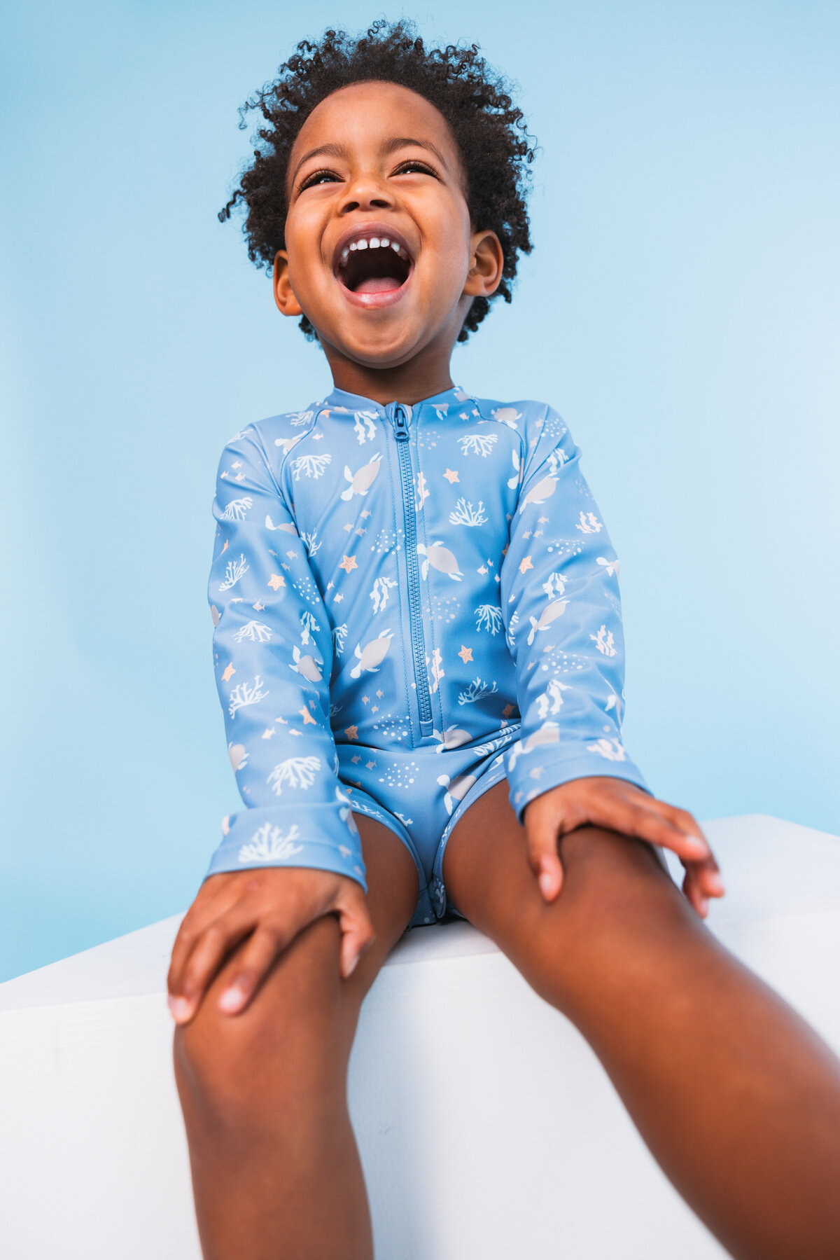 Joyful child laughing while wearing a long-sleeved blue Swim Essentials swimsuit featuring a sea turtle and coral print, seated against a light blue background.
