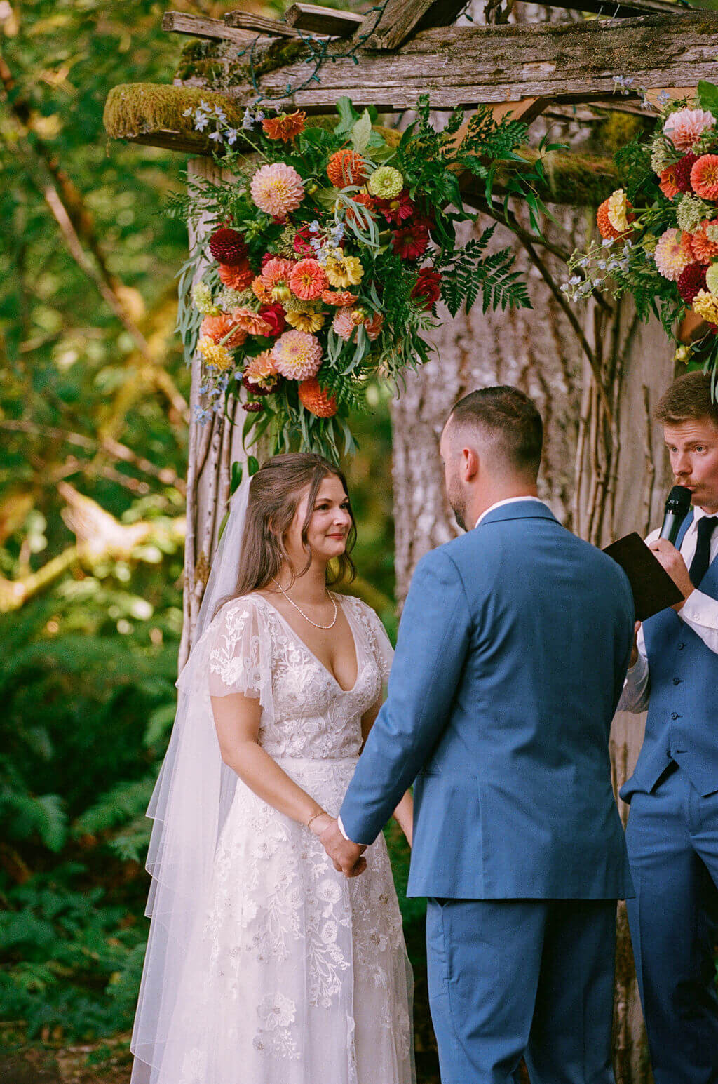 A bride and groom holds hands facing each other and standing under an arbor with a flower installation.
