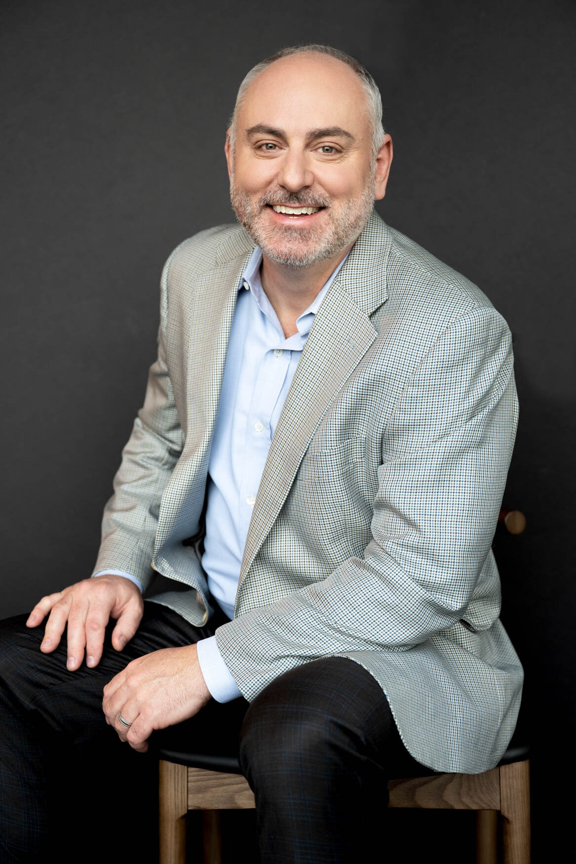 Professional headshot of a smiling man seated against a dark background, wearing a light gray blazer, light blue shirt, and dark slacks, exuding confidence and approachability.