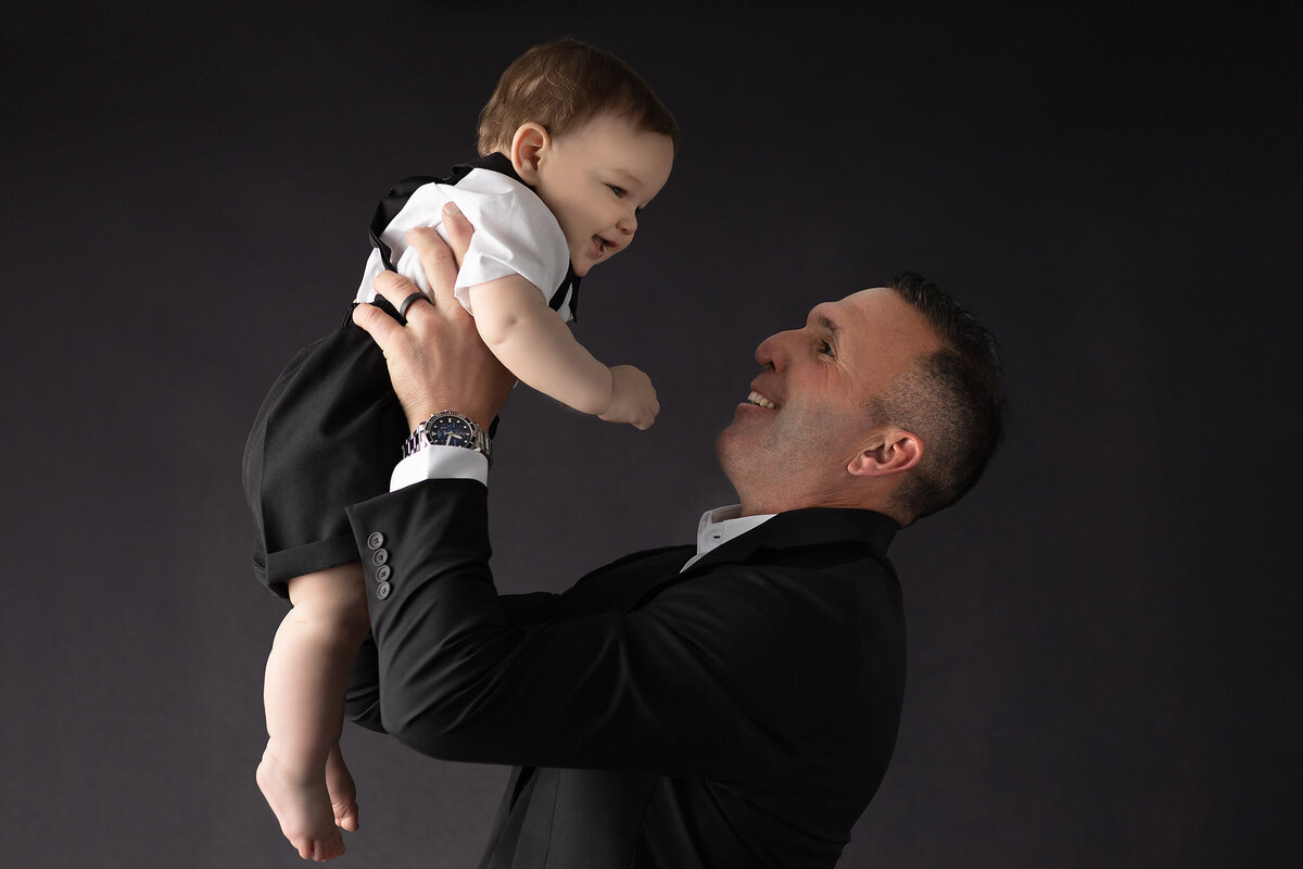 Dallas milestone photographer captures a joyful moment of a dad lifting his baby boy in the air, both dressed in formal black and white outfits against a dark background.