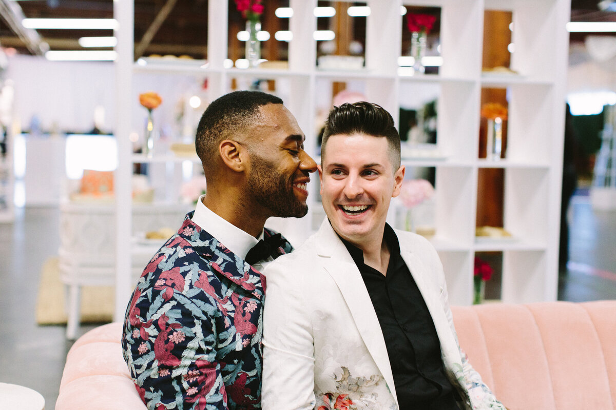 Two grooms posing together on their wedding day in Dallas, Texas. They sitting close to each other and smiling on a light pink couch in front of many shelves holding small bouquets. The groom on the left is wearing a blue jacket with intricate colorful floral patterns and a bowtie. The groom on the right is wearing a white jacket with intricate patterns on the bottom and a black dress shirt.