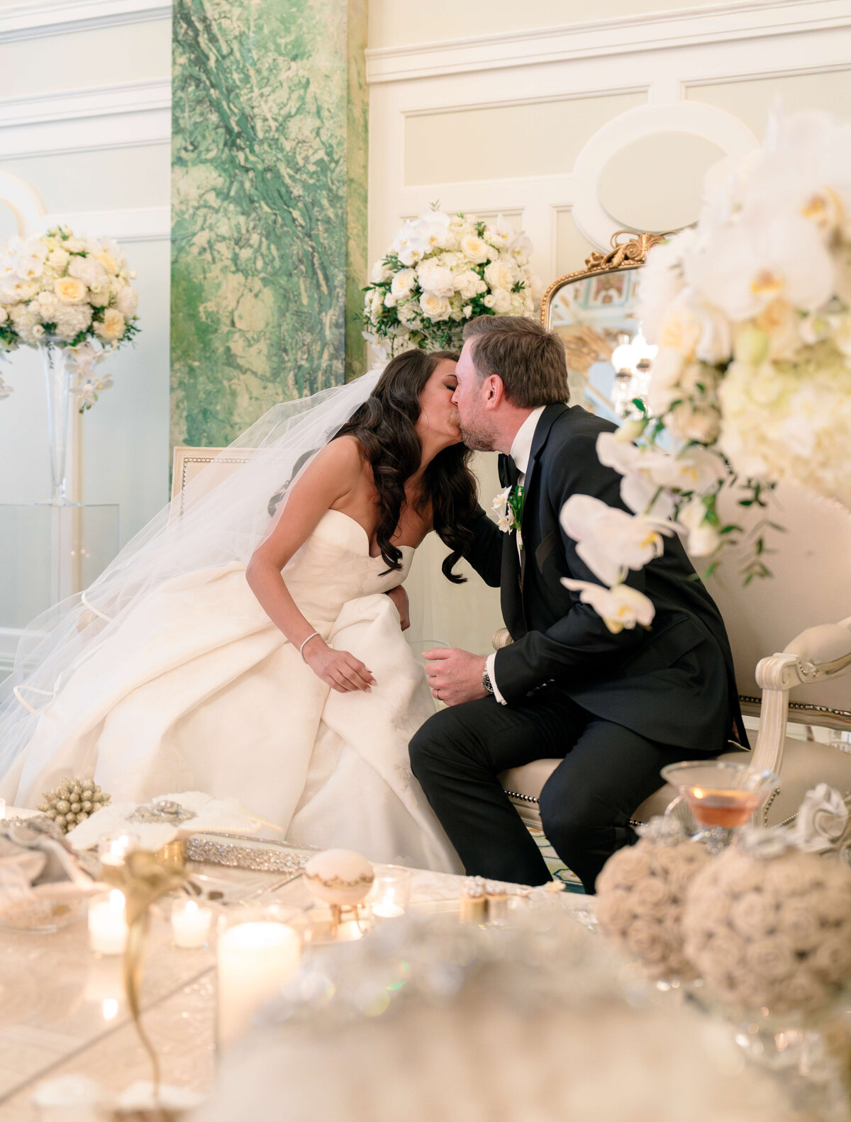 A bride and groom kiss while seated in an elegantly decorated wedding venue. They are surrounded by white flowers, candles, and soft lighting, with a lush green backdrop enhancing the romantic atmosphere.