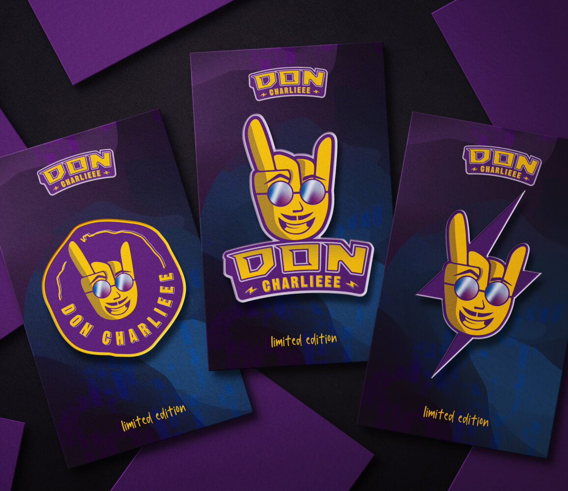 An array of enamel pins with three designs on the forefront: rock hand symbol with a smiling face wearing reflective sunglasses. The brand name is Don Charlieee.