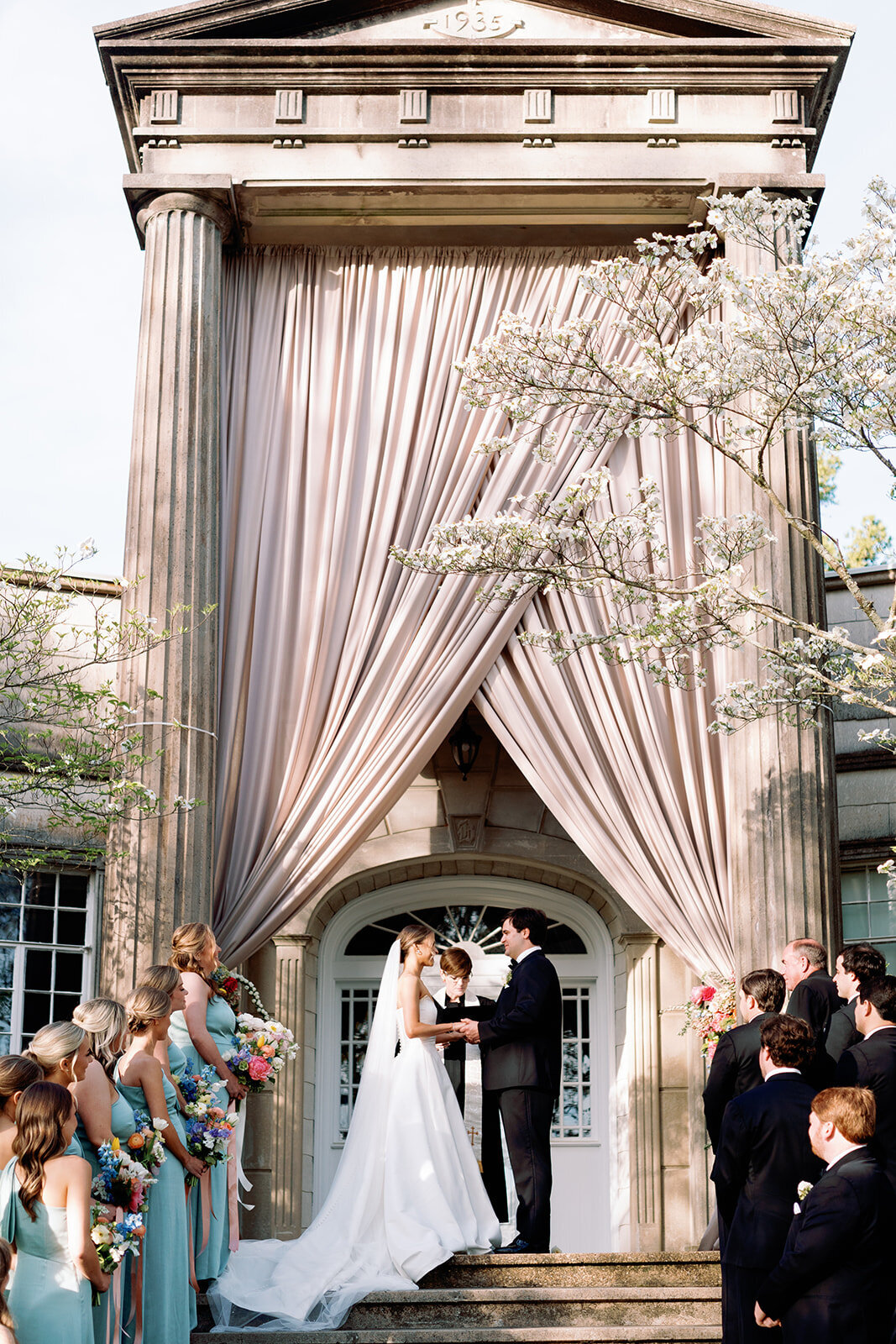 Bride and groom standing at the altar in front of the grand columned entrance of Burritt on the Mountain, surrounded by their wedding party during the ceremony in Huntsville, Alabama, photographed by Perry Hancock.
