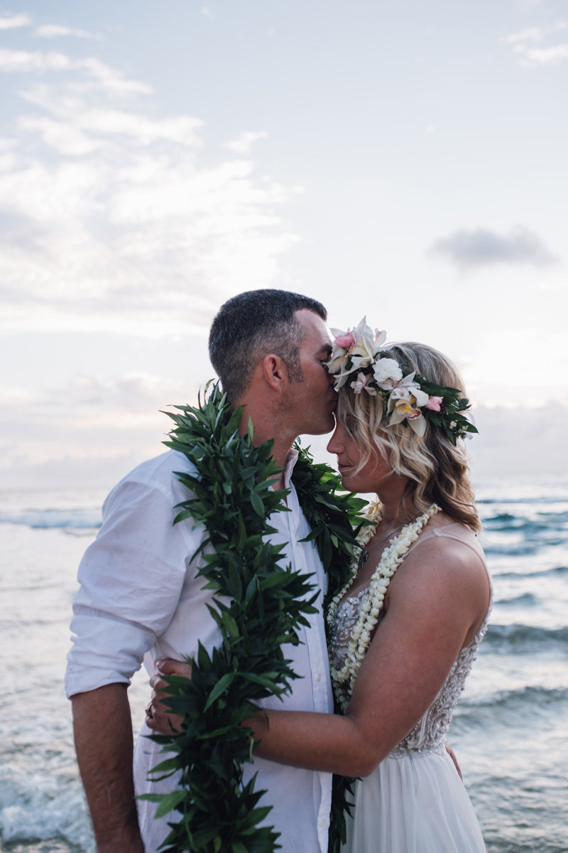 groom kissing brides forehead by the ocean with a male lei and she is wearing a haku lei
