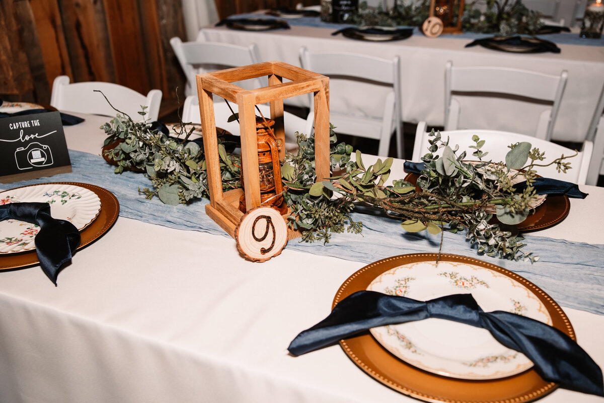 Details of wooden wedding reception center piece with greenery and blue napkins