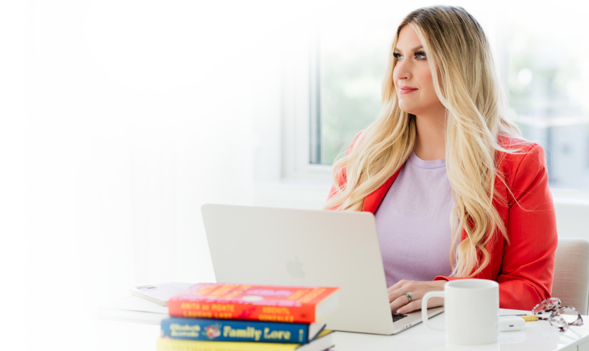 Headshot of Brittany Kues, podcast growth coach and owner of Kues media, sitting behind a desk. On the desk is a stack of colorful books that compliment her bold outfit, a coffee cup, and an open laptop.