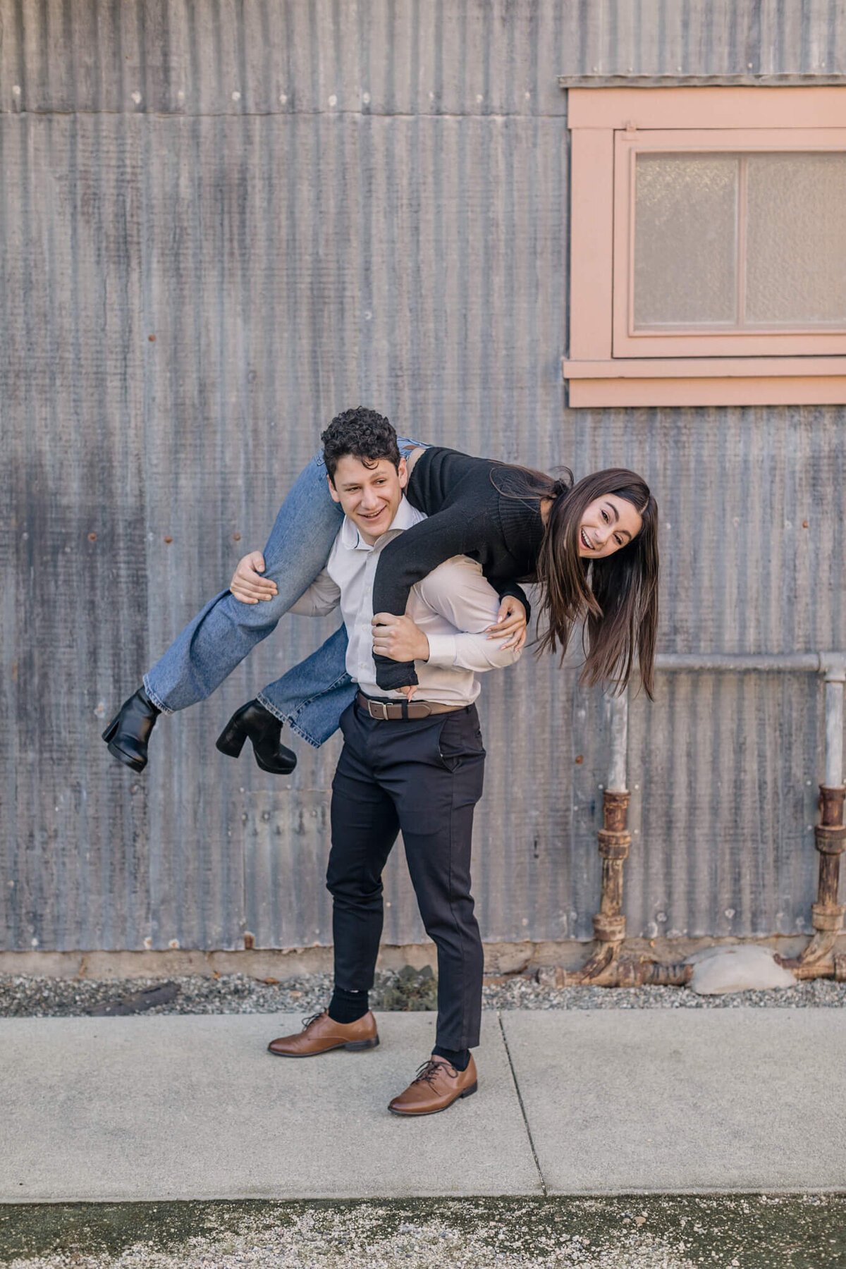 A brother with black hair wearing a grey button up shirt and navy dress pants holding his sister with long brown hair over his shoulders, her feet and head hanging off both of his shoulders as she's laughing at the camera.