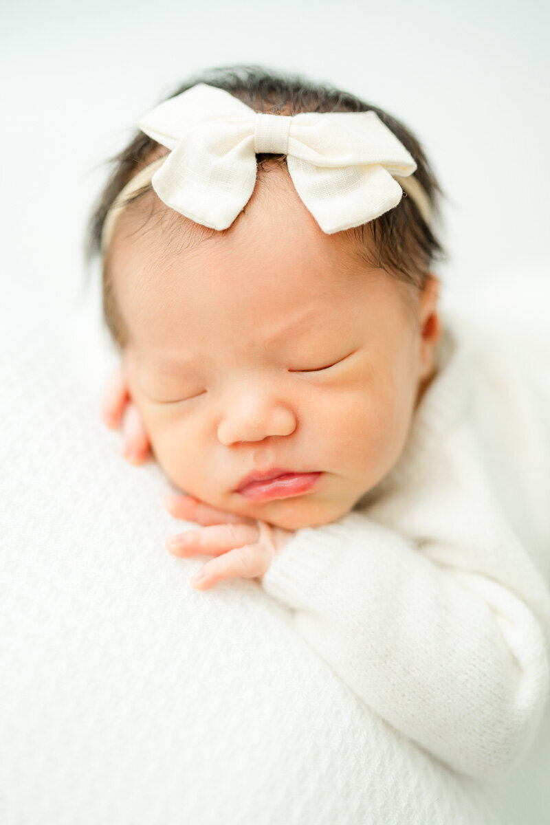 a newborn girl with a white bow in her hair sleeps on her stomach with her hands tucked under her chin during her posed newborn session in Austin. 
