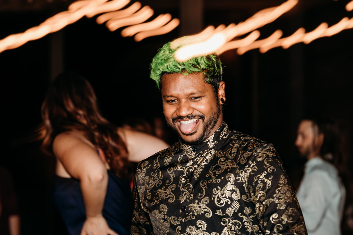 Man with green hair and a patterned shirt smiling at a wedding reception with warm lights streaking in the background