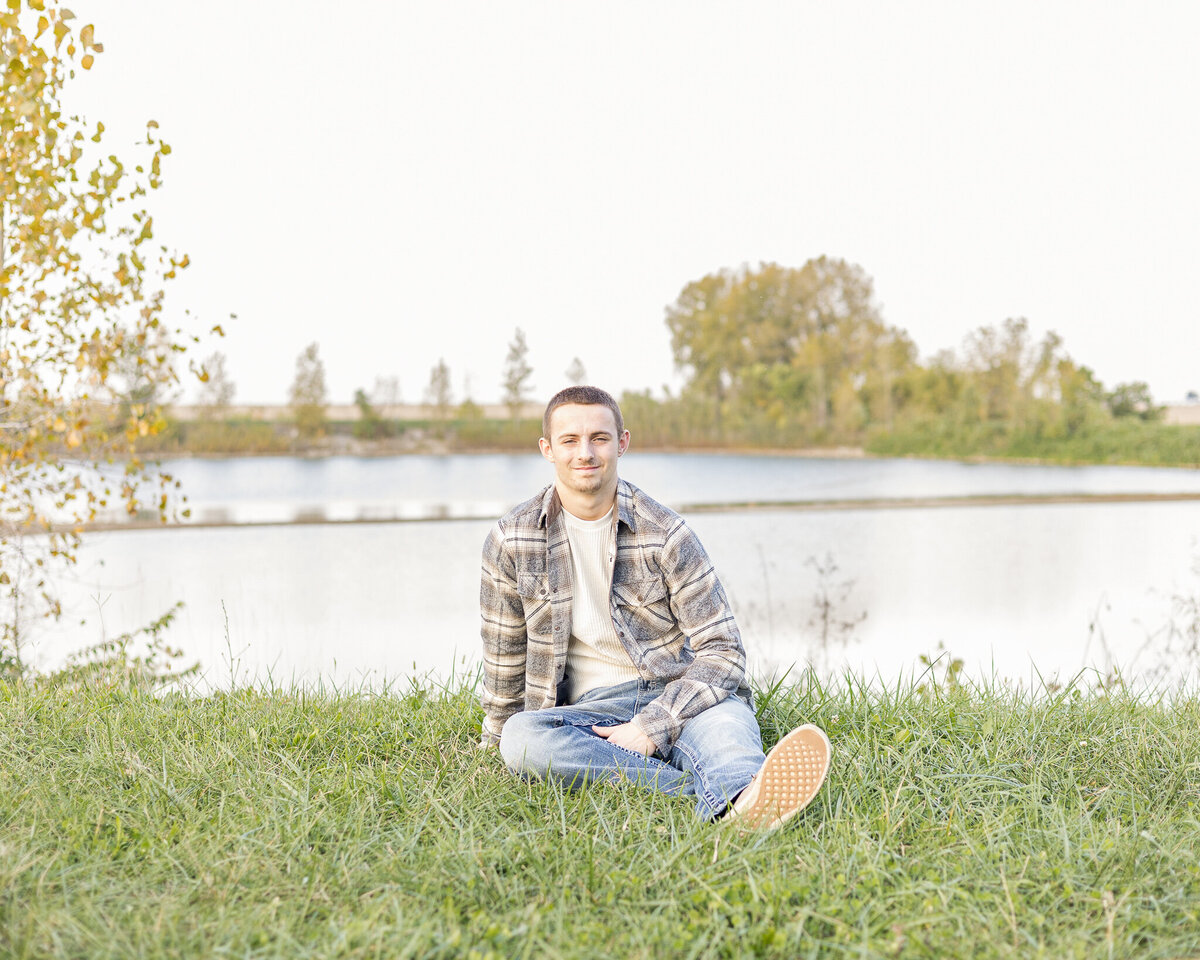 high school senior male sitting in grass with a pond and trees  behind him, wearing blue jeans, light tan shirt, blue jeans, looking at the camera softly smiling.