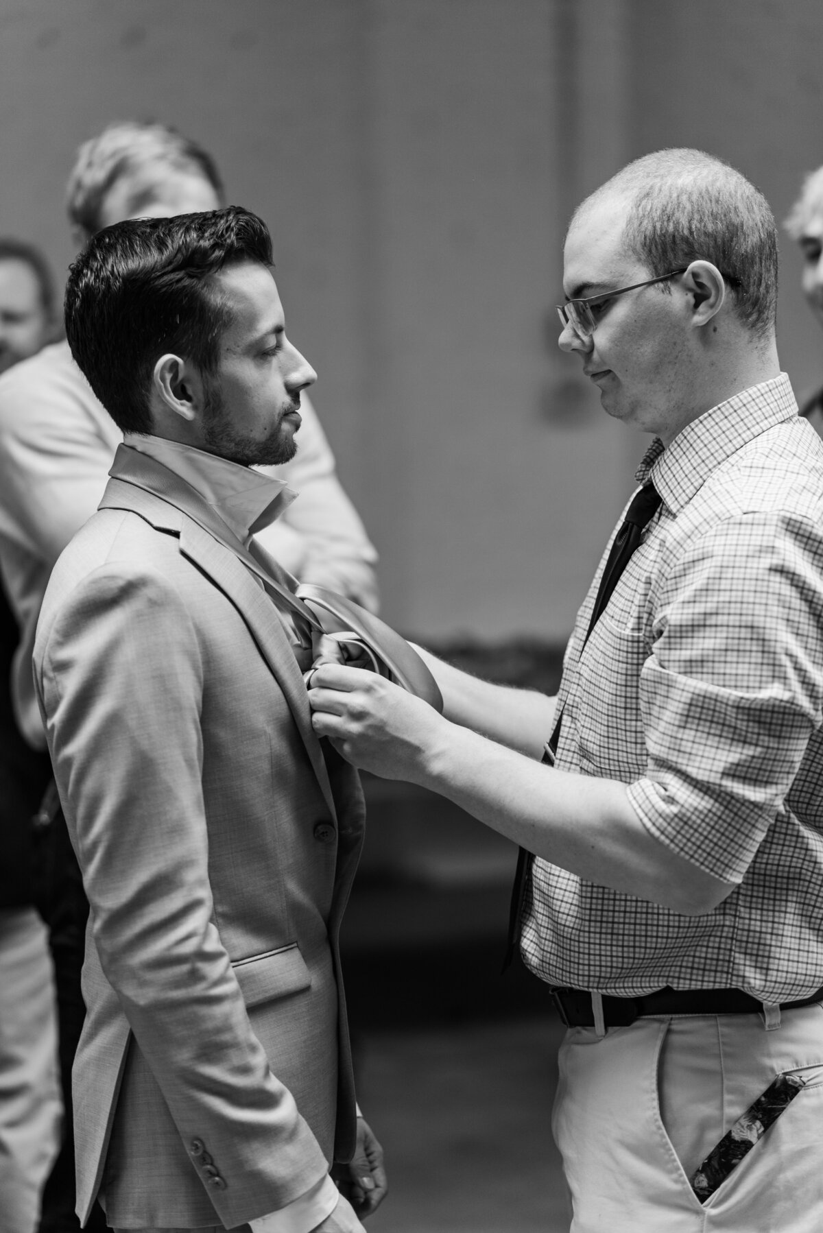 A black and white photo of a groomsman helping a groom tie his tie before the wedding ceremony at Skylight venue