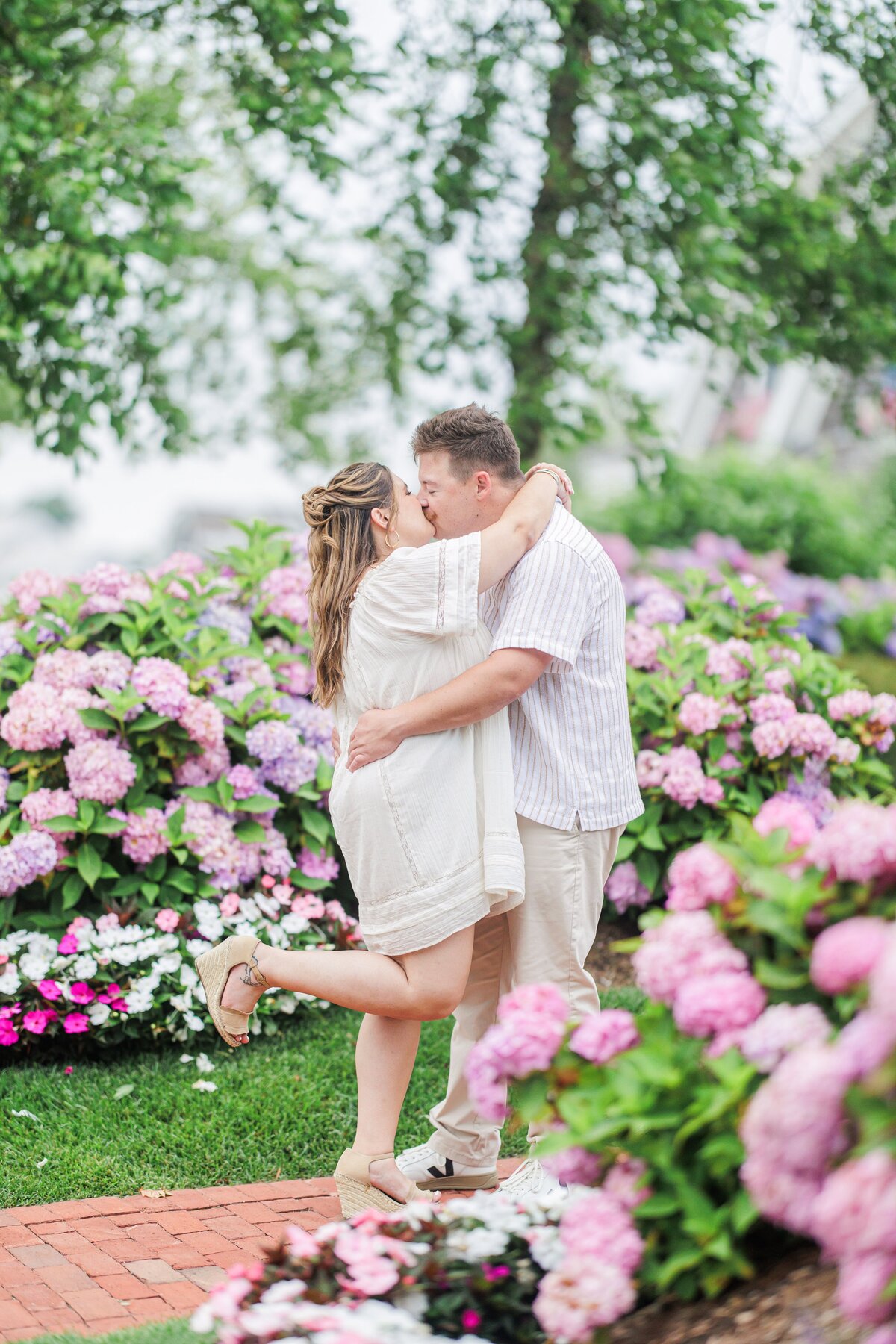 Couple kissing, the woman lifting one leg playfully, surrounded by blooming hydrangeas and garden flowers at Chatham Bars Inn in Chatham, Massachusetts — representing the portfolio of a Cape Cod engagement photographer.