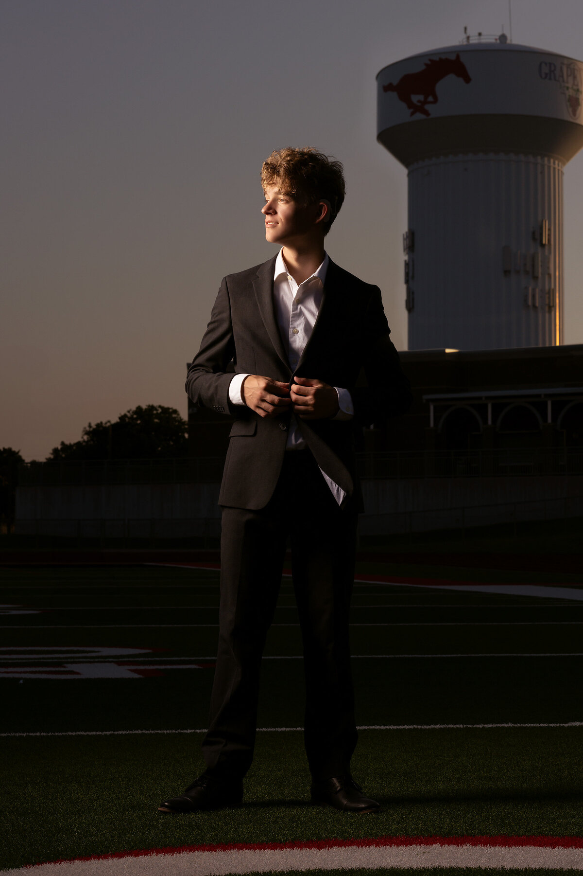 A senior guy stands confidently on the football field at Grapevine High School, adjusting his suit jacket. The iconic Grapevine water tower and the vibrant football lines create a striking backdrop in this Dallas senior photographer session.