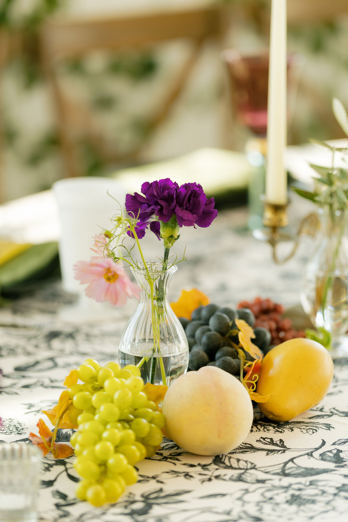 A lush wedding tablescape centerpiece designed by a fine-art wedding florist, featuring vibrant purple florals, blush pink cosmos, and delicate greenery arranged in a small glass bud vase. Surrounding the flowers are clusters of fresh grapes, golden apples, and soft blush peaches styled on a black-and-white floral linen, creating an organic, garden-inspired reception table design.