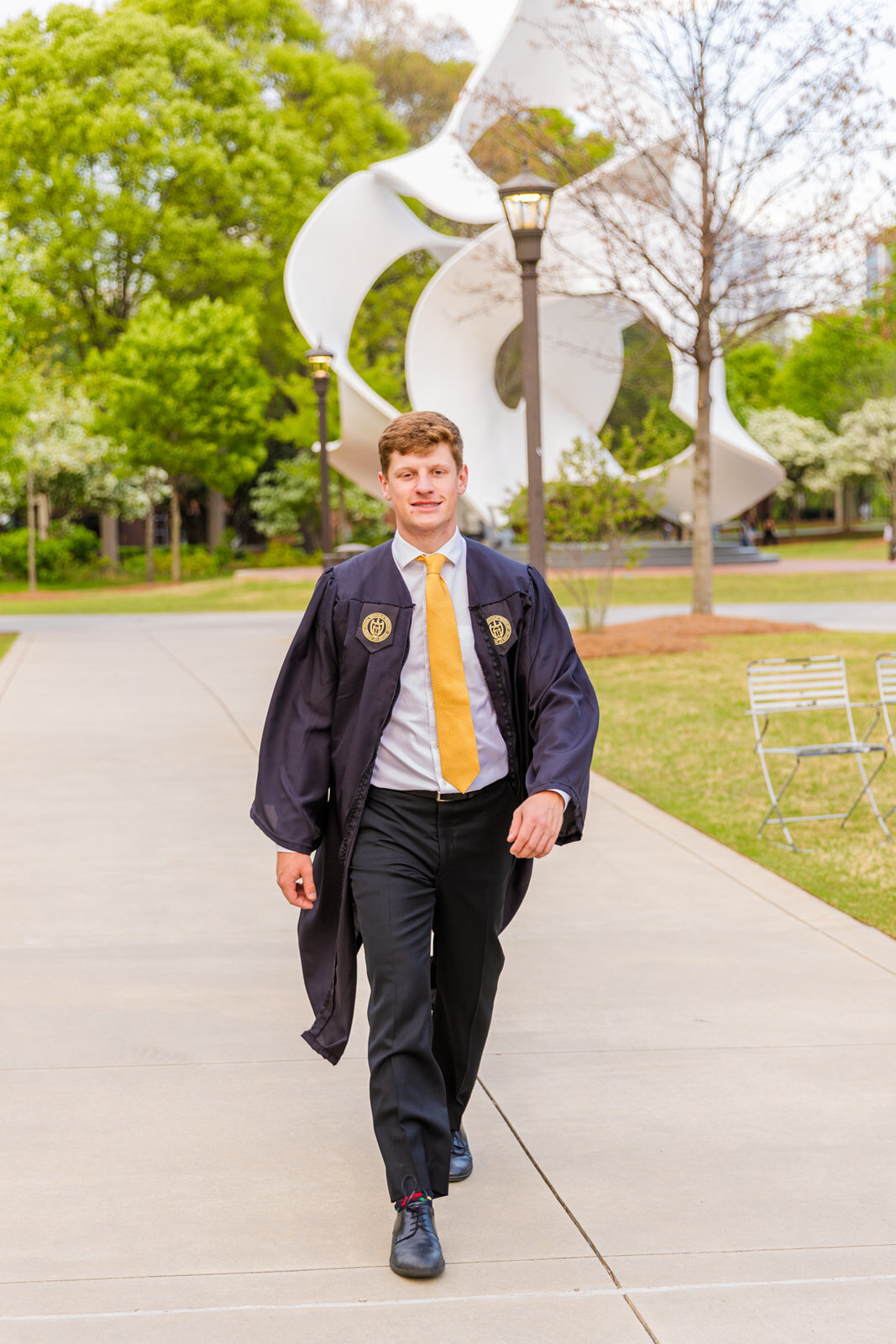 Undergrad senior boy walking outside on campus in front of a modern white statue wearing graduation gown over a white shirt and a yellow tie during graduation photo session with Atlanta photographer Laure Photography